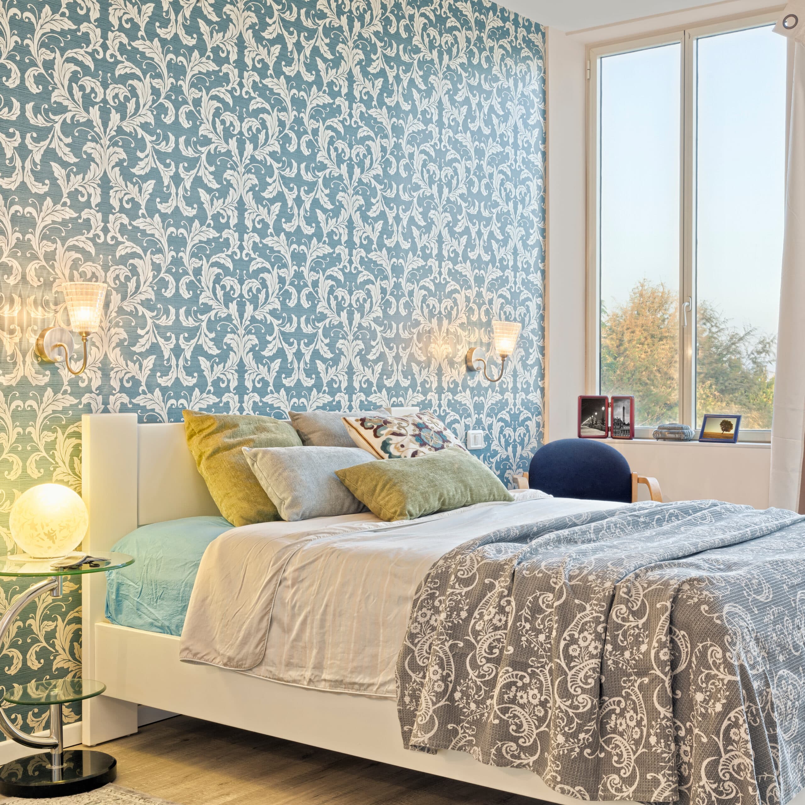 Chambre décorée avec papier peint bleu à motifs floraux blancs, lit blanc, coussins verts/gris, et lampe de chevet ronde lumineuse.