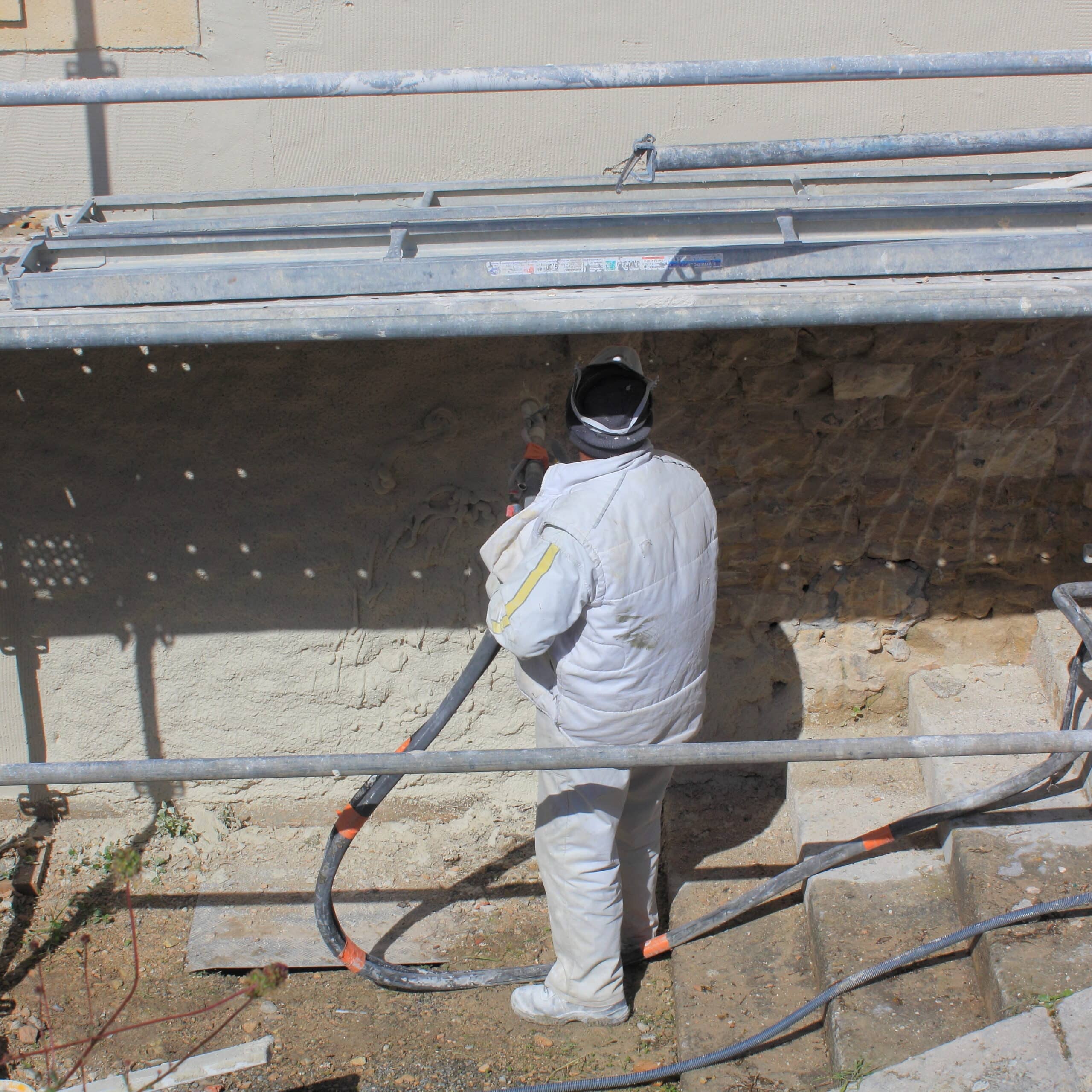 Ouvrier en combinaison blanche appliquant un enduit projeté sur un mur de pierre, sous un échafaudage.