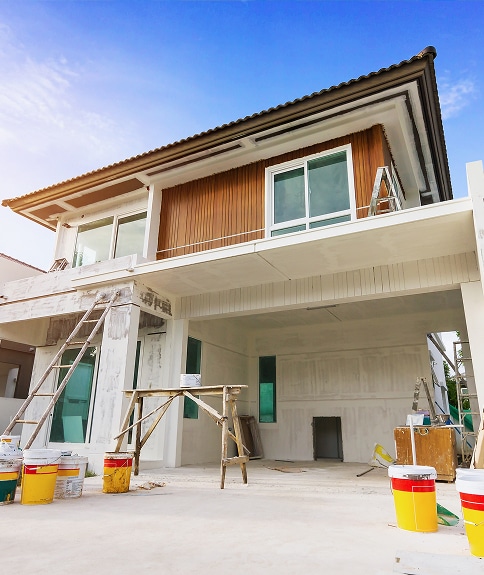 Maison moderne en construction avec revêtement bois et murs blancs, échelles et seaux de peinture jaunes au premier plan sous ciel bleu.