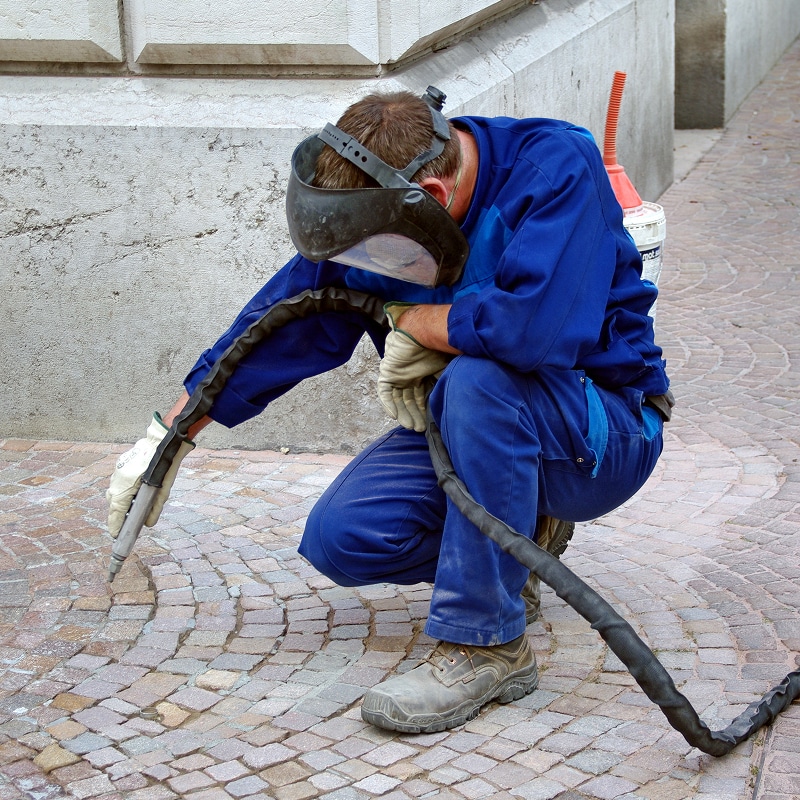 Ouvrier en tenue bleue accroupi, utilisant un pistolet de sablage sur des pavés devant un mur en pierre, portant un masque de soudure.