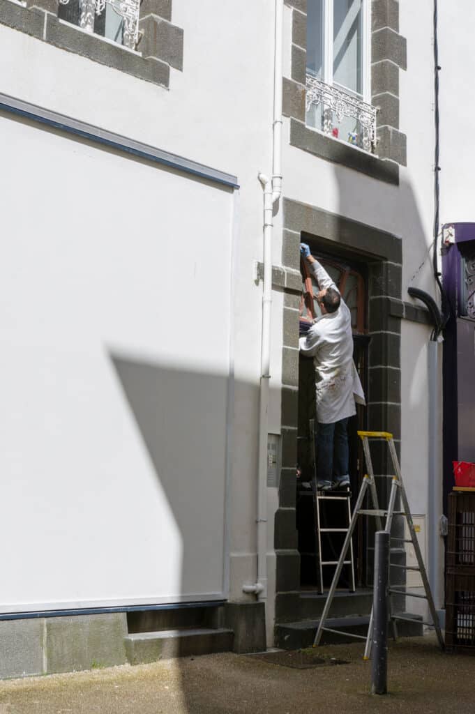 Peintre en blanc sur escabeau jaune, ajustant ou peignant l'encadrement d'une porte ou fenêtre d'une façade blanche avec pierres grises.
