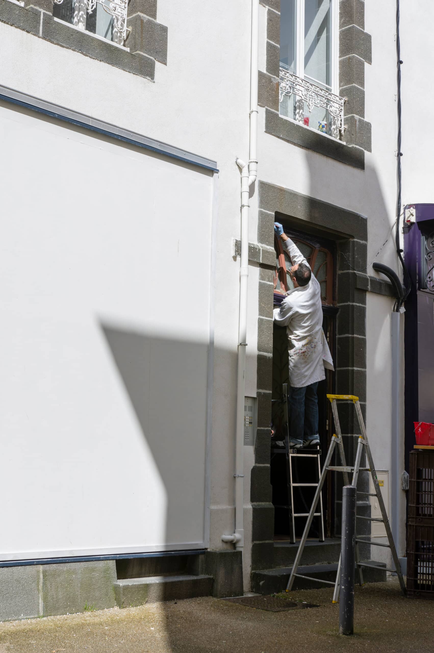 Peintre en blanc sur escabeau jaune, ajustant ou peignant l'encadrement d'une porte ou fenêtre d'une façade blanche avec pierres grises.